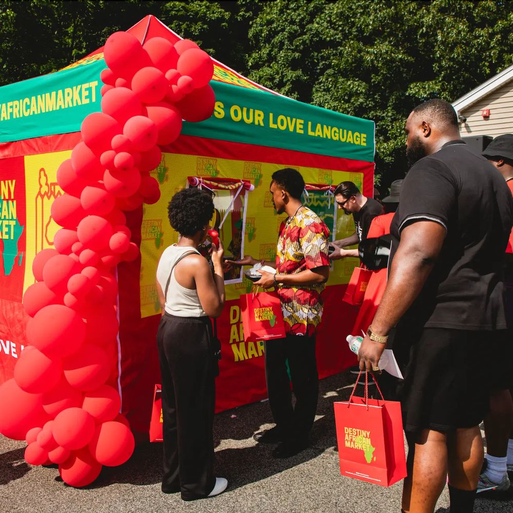 A branded Destiny African Market tent activation with a red balloon arch and a crowd of guests